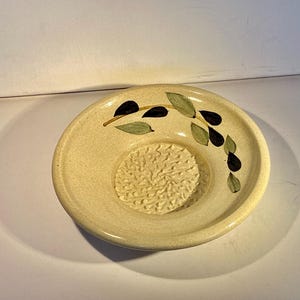 May include: A small, off-white ceramic bowl with a grater in the centre and a painted olive branch design. The bowl is perfect for grating garlic or ginger.