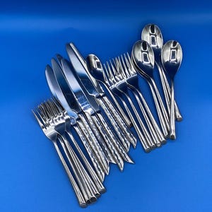 May include: A collection of stainless steel flatware including several dinner forks, knives with serrated edges, and spoons. Each piece features a polished, reflective finish and a slender, tapered handle design, arranged in a cluster on a blue surface.