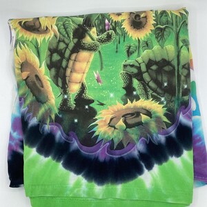 May include: A folded long-sleeve shirt with a vibrant tie-dye pattern. The shirt features a colourful graphic of turtles in a sunflower field with butterflies. The bottom of the shirt has a green and black tie-dye design.