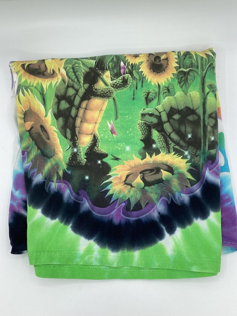 May include: A folded long-sleeve shirt with a vibrant tie-dye pattern. The shirt features a colourful graphic of turtles in a sunflower field with butterflies. The bottom of the shirt has a green and black tie-dye design.