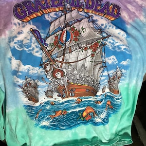 May include: Tie-dye t-shirt with a graphic of a ship sailing on the ocean, featuring skeletons and roses. The shirt has the words "Grateful Dead" across the top in purple and orange. The shirt's colors are blue, green, and purple.