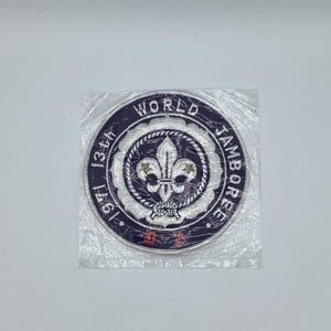 May include: A circular patch with the words "WORLD JAMBOREE" and "1971 13th" in white lettering on a dark blue background. The patch features a white fleur-de-lis emblem in the centre, surrounded by a white rope-like border. The patch is sealed in a clear plastic bag.