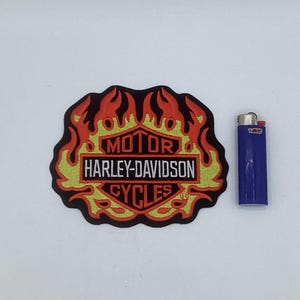 May include: A Harley-Davidson patch with a flame design in red, yellow, and black. The patch features the words "MOTOR HARLEY-DAVIDSON CYCLES" in a rectangular frame. A blue Bic lighter is next to the patch.