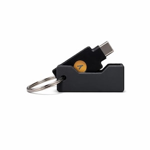 May include: A black USB-C adapter with a key ring attachment. The adapter has a rectangular base and a pivoting top section. A yellow circle with a white arrow is visible on the top section. The USB-C connector is silver.