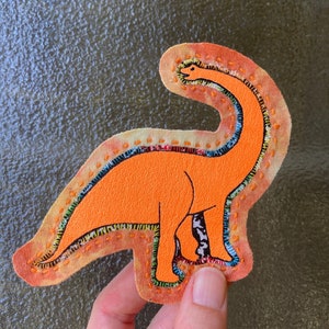 May include: A hand-sewn orange fabric dinosaur applique with colorful embroidery details. The dinosaur has a long neck and a small head with a smiling face.