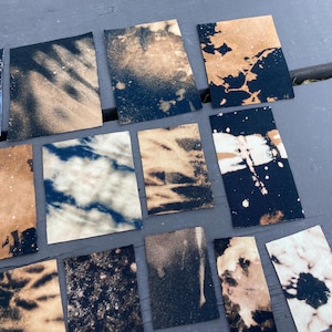 May include: A collection of 12 fabric squares with various bleach-splattered designs. The fabric is a dark blue color with light brown bleach splatters.