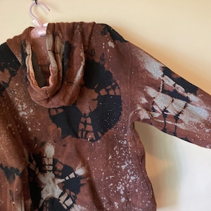 May include: A brown hoodie with a bleach tie-dye pattern in black and white. The hoodie has a hood and a zipper.