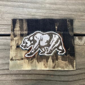 May include: A rectangular fabric patch featuring a dark, tie-dyed denim background with a central embroidered bear motif. The bear is depicted in shades of white and grey with a thick, textured outline, showcasing a detailed walking posture.