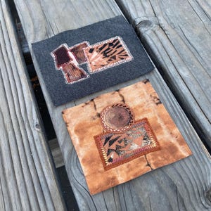 May include: Two rectangular fabric patches featuring intricate embroidery and appliqué. One patch is dark gray with brown and tan geometric shapes. The second patch is tan with a circular brown element and a rectangular section with dark patterns.