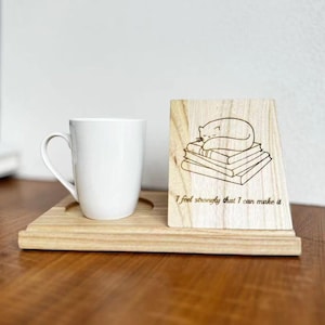 Puede incluir: Una taza de cerámica blanca y un posavasos de madera con un letrero de madera a juego. El letrero presenta una ilustración de un gato durmiendo sobre una pila de libros y el texto "I feel strongly that I can make it."