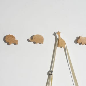 May include: Wooden animal-shaped wall hooks. The set includes a lion, elephant, giraffe, and hippo. The hooks are made of light-coloured wood and are mounted on a white wall. A beige strap hangs from the giraffe hook.