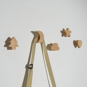 Puede incluir: Ganchos de pared de madera con forma de árbol, luna, nube, estrella y corazón. Los ganchos son de madera clara y están montados en una pared blanca. Una bolsa beige cuelga del gancho en forma de luna.
