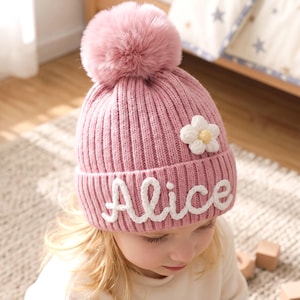 Op de afbeelding: Een oudroze geribbelde gebreide muts met een grote bijpassende pompon van imitatiebont bovenop. De muts is versierd met een witte gehaakte bloemapplicatie en de naam Alice geborduurd in witte schrijfletters op de omgeslagen rand.
