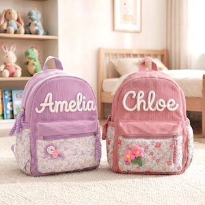 Op de afbeelding: Twee rugzakken van corduroy, één in lavendel en één in oudroze, elk met een voorvak met bloemenpatroon. De lavendelkleurige tas heeft de naam Amelia in witte letters en de roze tas heeft de naam Chloe in witte letters met bloemaccenten.