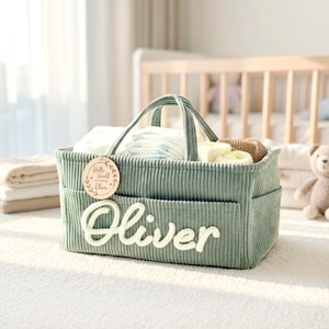 May include: A sage green corduroy baby caddy with the name "Oliver" in white script. The caddy has a handle and pockets, filled with diapers and baby items. A wooden tag reads "Hello World, Welcome Oliver."
