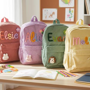 Może przedstawiać: Cztery sztruksowe plecaki w kolorze różowym, fioletowym, zielonym i żółtym, każdy z imieniem wyhaftowanym z przodu. Plecaki mają przednią kieszeń i małą aplikację ze zwierzątkiem lub kwiatkiem. Artykuły szkolne na stole.