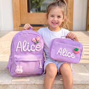 May include: A lavender corduroy backpack and a matching rectangular lunch bag, both featuring the name Alice in white script lettering. The backpack is decorated with two pink flowers and a small pink bunny, while the lunch bag has a strawberry patch.