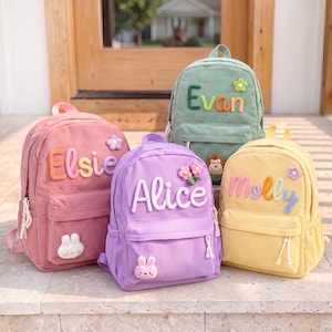 Könnte beinhalten: Vier Rucksäcke aus Kord in Rosa, Lila, Gelb und Grün, jeweils mit einem personalisierten Namen in erhabenen Buchstaben, einem kleinen Blumenakzent und einem niedlichen Tieraufnäher auf der Vordertasche, darunter ein weißer Hase, ein Igel und ein rosa Hase.