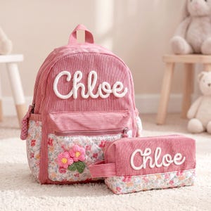 Peut inclure: Un sac à dos en velours côtelé rose poudré et une trousse assortie, tous deux ornés du prénom Chloe en lettres de corde blanche. La poche avant du sac et la base de la trousse présentent un motif floral avec deux fleurs roses crochetées.