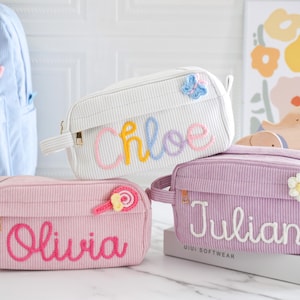 Puede incluir: Tres estuches de lápices de pana en rosa, blanco y lavanda, cada uno con un nombre bordado con hilo de colores. El estuche rosa tiene un aplique de piruleta, el estuche blanco tiene una mariposa y el estuche lavanda tiene una flor.