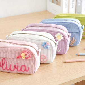 Puede incluir: Estuches de lápices de pana en colores pastel: rosa, blanco, lavanda, azul y verde. Cada estuche tiene cierre de cremallera y detalles bordados, incluyendo nombres y adornos florales o de caramelos. Hay lápices cerca.