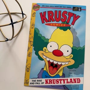 Könnte beinhalten: Ein Comic-Heft mit dem Titel "Krusty Comics" mit der Figur Krusty dem Clown. Das Cover hat einen blauen Hintergrund mit dem Titel in Rot. Der Clown hat gelbe Haut, grüne Haare und eine rote Nase. Der Text "THE RISE AND FALL OF KRUSTYLAND" ist ebenfalls sichtbar.