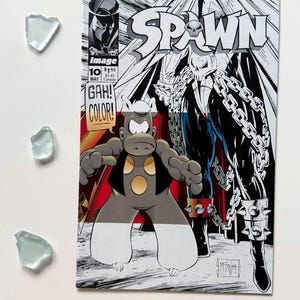 May include: A comic book cover for "Spawn" issue 10, featuring a black and white illustration of a skull-faced character with chains and a cape. The cover also includes a cartoon hippo character and the text "GAH! COLOR!"