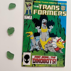 May include: A vintage comic book titled "The Transformers" with a robot dinosaur illustration on a green and blue background. The comic book has the text "AND NOW... THE DINOBOTS!" and "MARVEL" on the cover. Three pieces of green sea glass are also present.