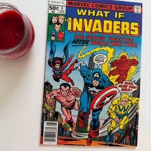 Puede incluir: Un cómic vintage titulado "What If The Invaders Had Stayed Together After World War Two?" presenta al Capitán América y otros superhéroes. La portada es vibrante con colores primarios y poses de acción. Un tarro de mermelada roja está en el fondo.