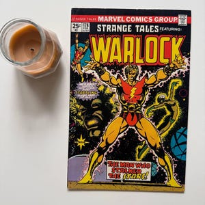 May include: A vintage comic book titled "Strange Tales" featuring "Warlock." The cover art displays a superhero in a red and yellow costume with the text "The Man Who Stalked the Stars!" A lit candle in a glass jar is in the upper left corner.