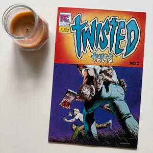 May include: A vintage comic book titled "Twisted Tales" No. 2, featuring a horror-themed illustration with a figure holding an axe. The cover has a red and orange background with blue text. A lit candle in a glass jar is in the upper left corner.