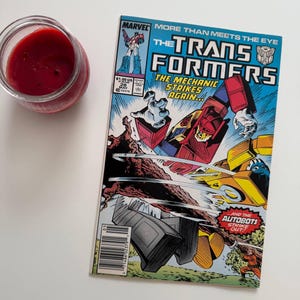 May include: A vintage comic book titled "The Transformers" with a vibrant cover featuring robot characters in action. The comic book has a blue background with red, yellow, and gray robots. A red candle in a glass jar is in the upper left corner.