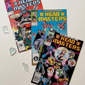 May include: Three vintage Marvel comic books from the Transformers: Headmasters series. The covers feature colourful illustrations of robots and characters, with the Marvel logo and issue numbers visible. The comics are in good condition.