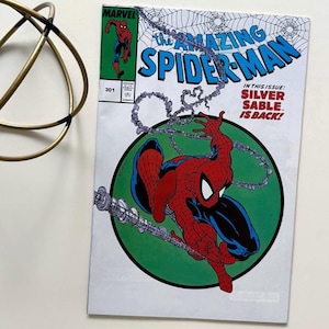 May include: The Amazing Spider-Man comic book cover featuring Spider-Man in a red and blue suit, swinging from a web. The cover has a green circle with the text "Silver Sable is Back!" and the Marvel logo.