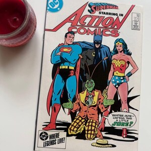 Puede incluir: Una portada de cómic con Superman, Batman y Wonder Woman. La portada es blanca con un diseño rojo, azul y amarillo. El título del cómic es "Action Comics" y el texto "Superman starring in" está por encima del título. El texto "Where Legends Live!" está en la esquina inferior izquierda. El texto "Smatter, guys - can'tcha take a little joke?" está en un globo de diálogo en la esquina inferior derecha.