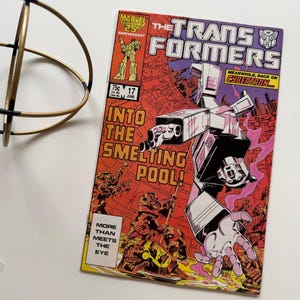 May include: A vintage Transformers comic book. The cover is predominantly red, with a large robot arm and the title "The Transformers." Text includes "Into the Smelting Pool!" and "More Than Meets The Eye."