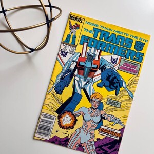 May include: A comic book cover featuring the Transformers, a group of robots who can transform into vehicles. The cover is yellow with a blue and red robot in the centre. The text on the cover reads "More Than Meets The Eye" and "The Transformers".