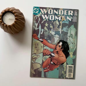 May include: A bronze-colored, geometric candle holder with a white candle next to a Wonder Woman comic book. The comic book cover features Wonder Woman in a dynamic pose, with the title "Wonder Women" and issue number 174.