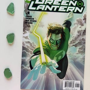 Puede incluir: Un cómic de "Green Lantern" con una ilustración de superhéroe en verde y blanco. La portada presenta al personaje de Green Lantern con un anillo brillante y el logotipo de DC Comics. Tres trozos de vidrio de mar verde están a la izquierda.