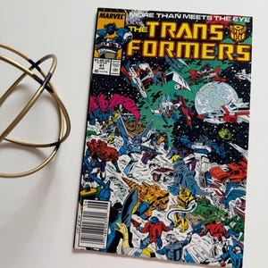 May include: A vintage comic book titled "The Transformers" with vibrant illustrations of robots in action. The cover features the Marvel logo and the text "More Than Meets The Eye". The comic is set against a white background.