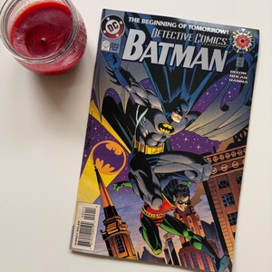 May include: A vintage Batman comic book featuring Batman and Robin, with the text "DETECTIVE COMICS" and "BATMAN". A red candle in a glass jar is in the upper left corner. The comic book is on a white surface.