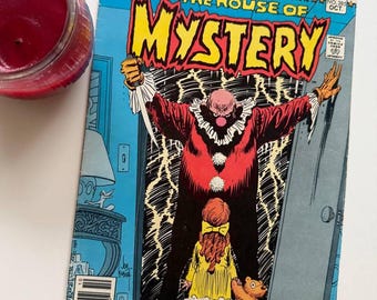 Peut inclure: Une bande dessinée vintage intitulée "The House of Mystery" avec un clown menaçant, une jeune fille et un ours en peluche. La couverture est bleue, rouge et jaune, avec le logo DC Comics. Une bougie rouge est visible. Mesures non applicables.