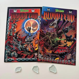 May include: Two comic books titled "Spawn Bloodfeud" with vibrant covers featuring dark fantasy art. The covers display a variety of skulls, chains, and monstrous figures in shades of red, orange, and purple. Three pieces of sea glass are at the bottom.