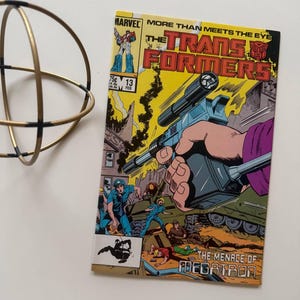 May include: A vintage comic book titled "The Transformers" with a vibrant cover featuring a robot holding a large weapon. The comic has a yellow and orange background with the text "The Menace of Megatron". A gold metal decorative sphere is in the upper left corner.