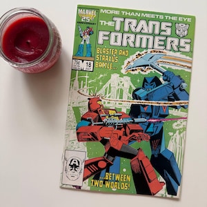 May include: A vintage "The Transformers" comic book with a green cover featuring robot illustrations. Beside it is a red candle in a glass jar. The comic book has the text "More Than Meets The Eye" and is a 25th Anniversary edition.