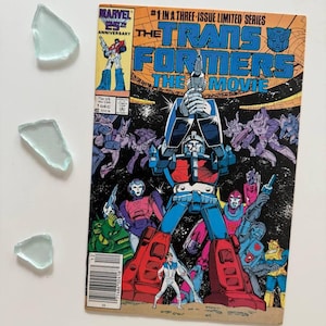 May include: A retro comic book cover for "The Transformers: The Movie" with a large robot in blue, red, and white. The cover includes the Marvel Comics logo and the text "#1 in a three-issue limited series". Several pieces of sea glass are in the image.