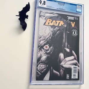 May include: A graded Batman #653 comic book, rated 9.8, in a protective case. The cover shows the Joker with a gun. A black bat decoration is placed near the comic book, evoking the Batman theme.