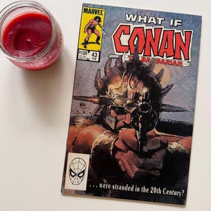 Puede incluir: Un cómic vintage de Marvel Comics "What If Conan the Barbarian", número 43, con una ilustración de portada de Conan sosteniendo un arma. Una vela roja en un tarro de cristal está a la izquierda del cómic. El cómic tiene un logo de Spider-Man.