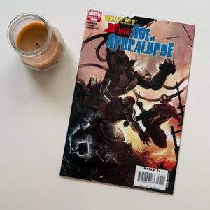 Puede incluir: Un cómic titulado "X-Men Age of Apocalypse" con ilustraciones dinámicas de superhéroes en acción. La portada presenta una escena de batalla con varios personajes en colores vibrantes. Una vela encendida en un tarro de cristal está al lado del cómic.