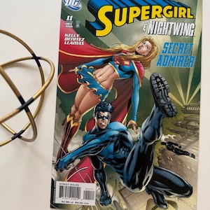 Può includere: Copertina di fumetto con Supergirl e Nightwing. L'illustrazione della copertina mostra Supergirl in un costume rosso e blu con un mantello rosso, e Nightwing in un costume blu con una maschera nera. Il titolo del fumetto è "Supergirl e Nightwing: Secret Admirer".